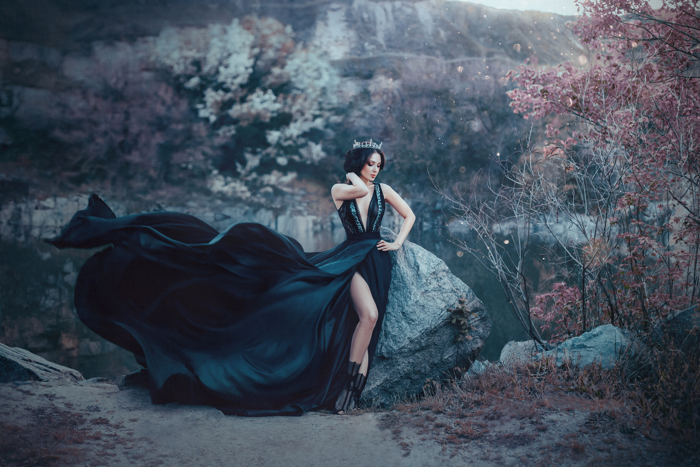 The dark queen pose against the background of gloomy rocks. A luxurious black dress with a long train fluttering in the wind denuding her leg. Elegant, collected hairstyle with a gothic crown